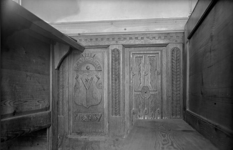 A black and white photo of a carved wooden panel showing a shield and foliate designs
